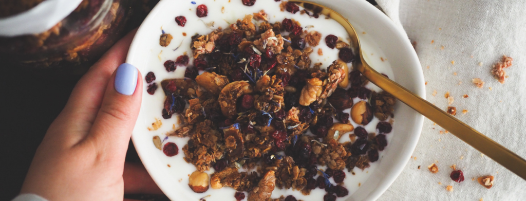 A bowl filled with creamy yogurt is topped with granola, coconut flakes, walnuts, cranberries, and other dried fruits and nuts. A hand with lavender nail polish gently holds the bowl. A gold spoon rests inside, while a few granola pieces are scattered on a cloth nearby.