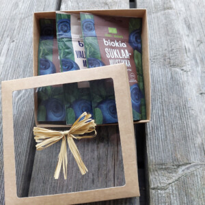 A "Gift box - chocolate bilberries" is displayed open on a wooden surface. Inside, three packages of chocolate bilberries are nestled together. A decorative straw ribbon drapes across the clear lid.