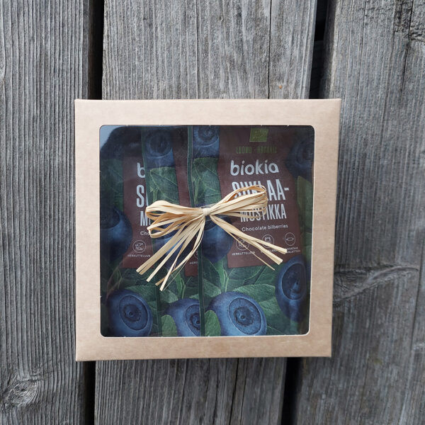 The Gift box - chocolate bilberries showcases two packs of chocolate-covered bilberries through its transparent window. Decorated with a rustic straw ribbon, this square cardboard box makes for an ideal chocolate-themed surprise. The wooden surface background enhances its charming presentation.