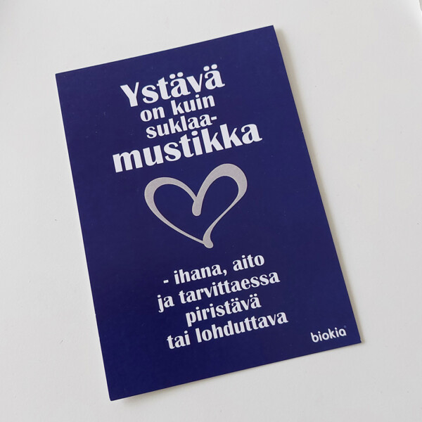 Finnish blue card with white text: "Ystävä on kuin suklaamustikka - ihana, aito ja tarvittaessa piristävä tai lohduttava," features a silver heart design like the Gift box - chocolate bilberries. The "biokia" logo is at the bottom.